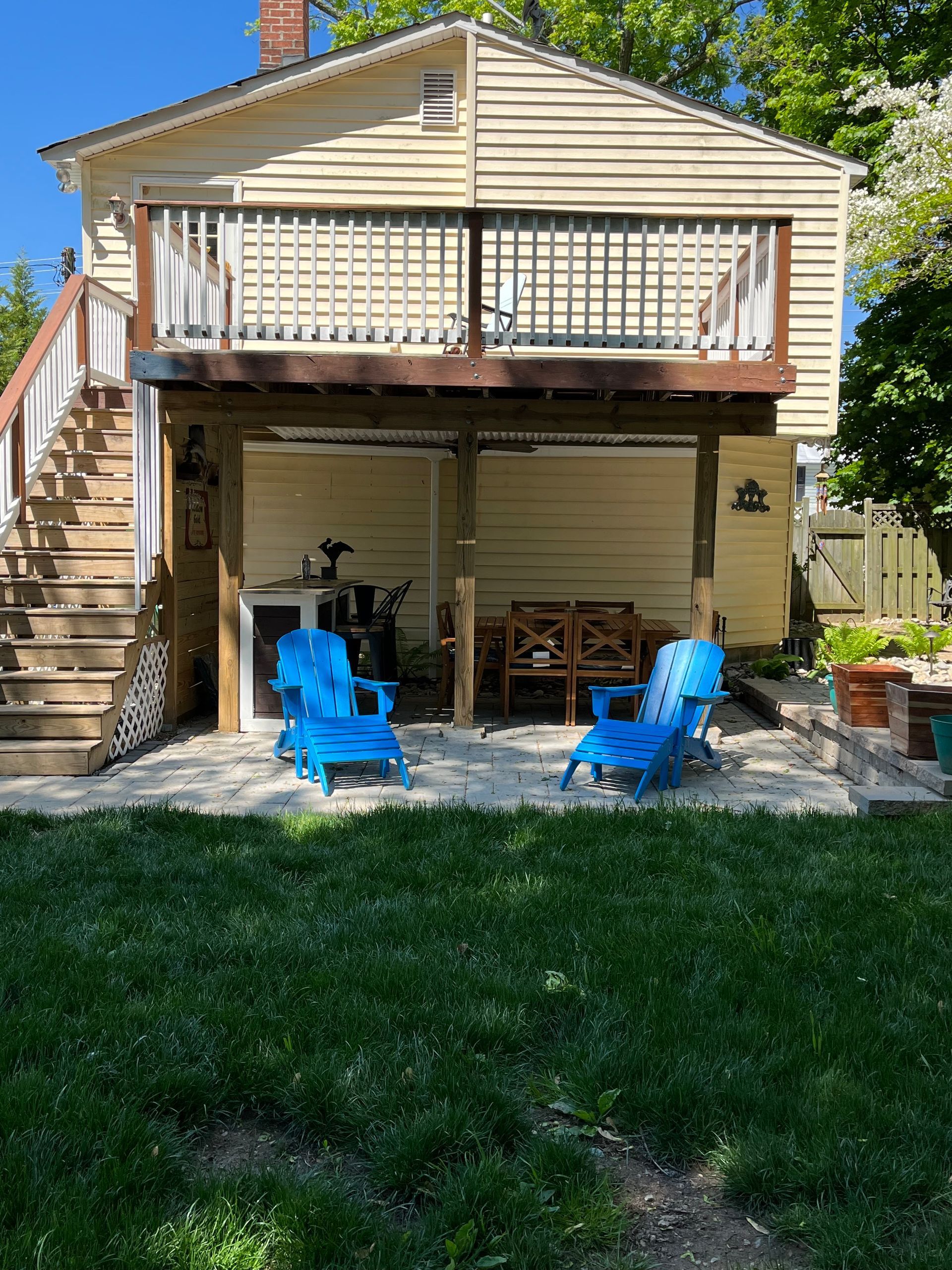 Two bright blue Adirondack chairs sit on a gravel patio under a wooden deck attached to a light yellow house.