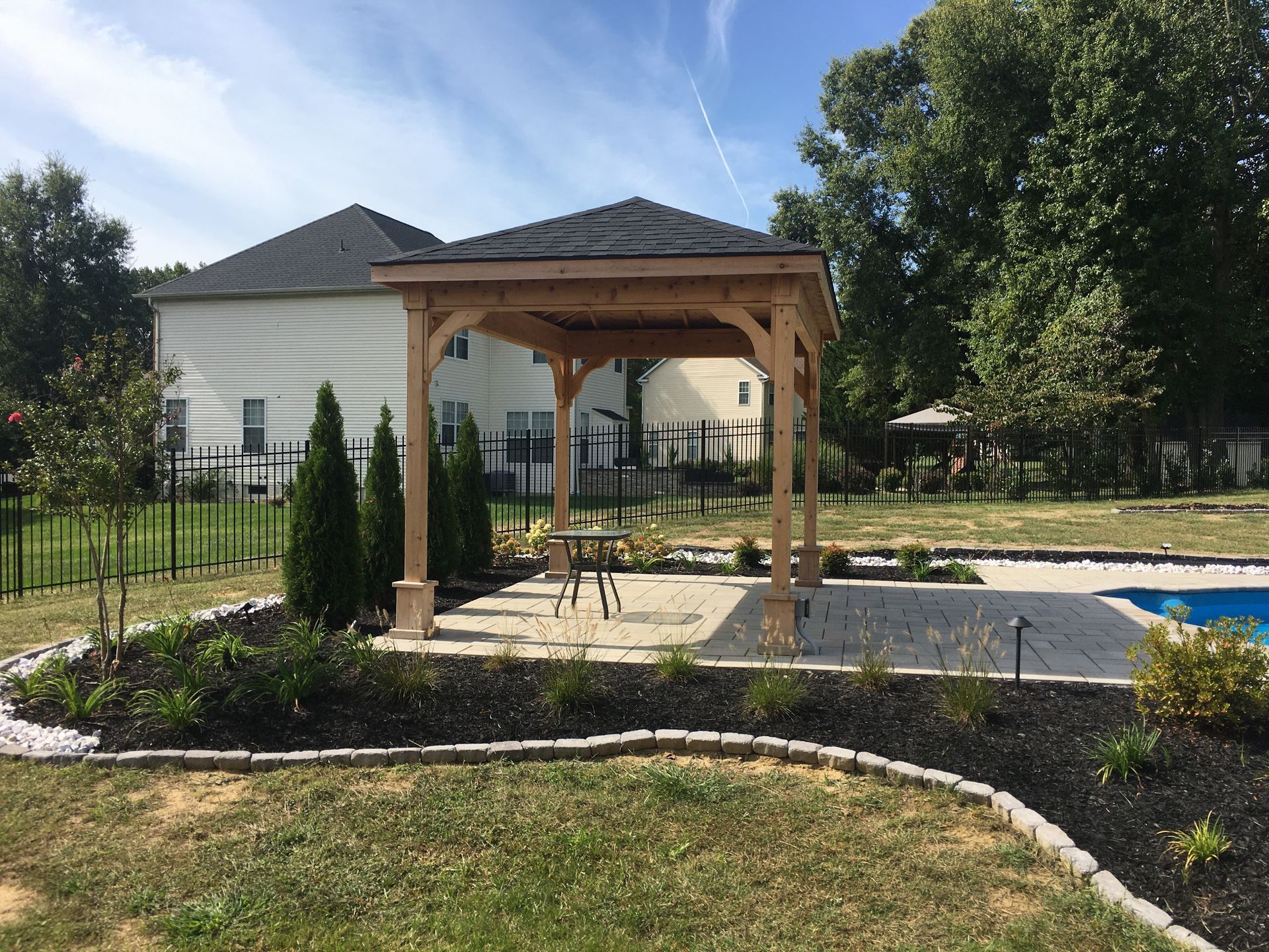 A wooden pergola stands on a paved patio beside a swimming pool, surrounded by mulched garden beds and a grassy lawn.