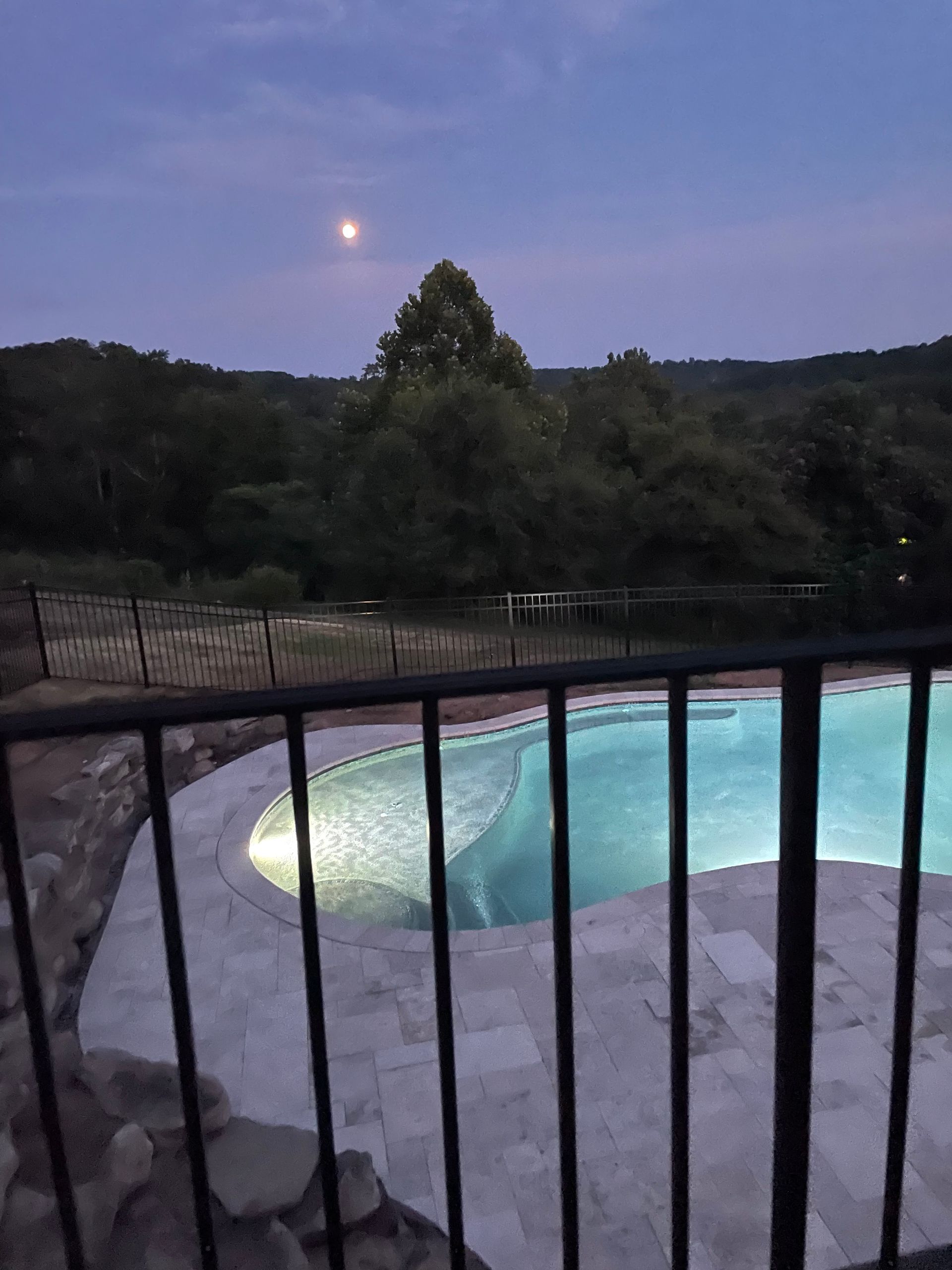 A glowing swimming pool surrounded by a stone patio at dusk, with a moon visible above silhouetted trees.