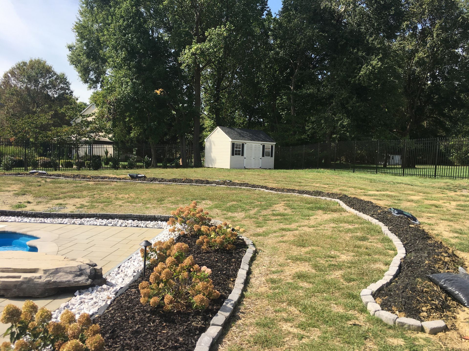 A landscaped backyard with a curved stone-edged flower bed, mulch, and small shrubs bordering a paved pool patio.