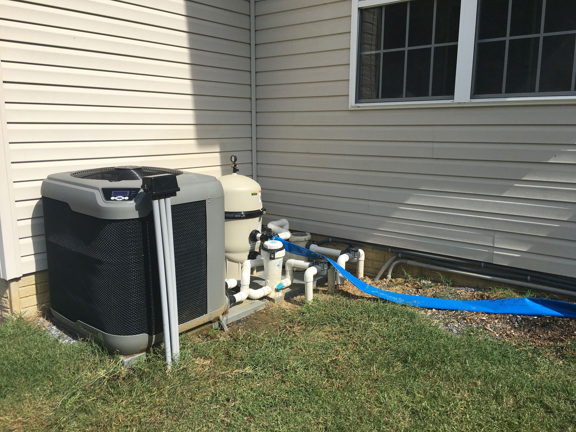 A pool filter system and an air conditioning unit sit on the grass next to a house with beige siding.