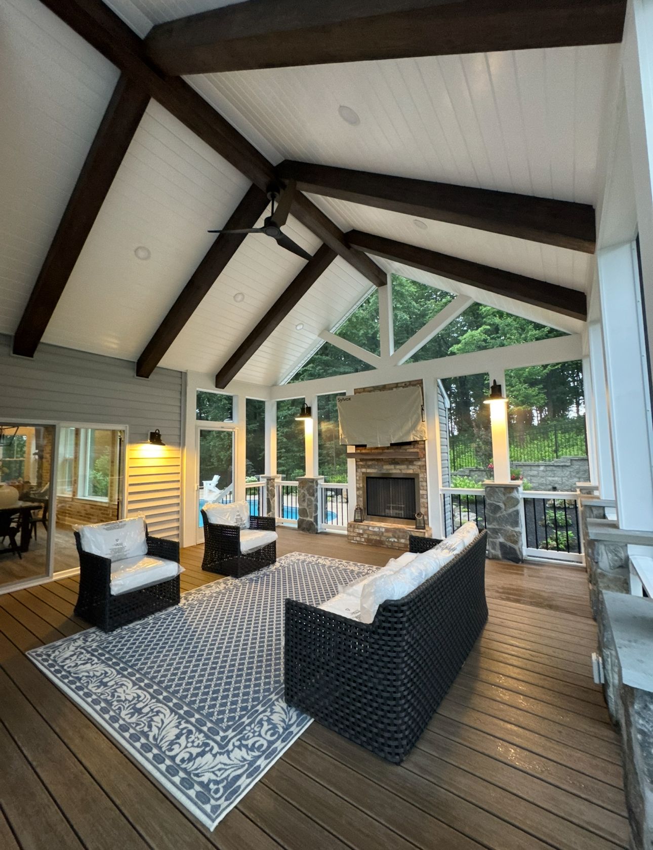 A furnished, covered patio with vaulted wood-beamed ceilings, a stone fireplace, and a large patterned area rug.