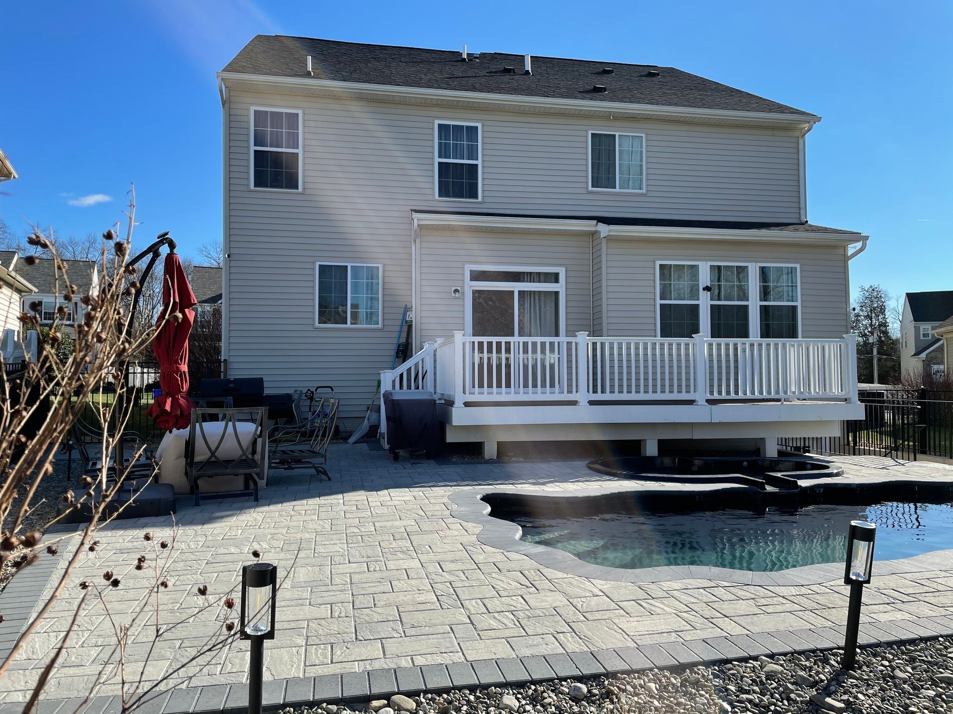 A two-story tan house with a white deck, patio, and swimming pool in a suburban backyard under a bright blue sky.