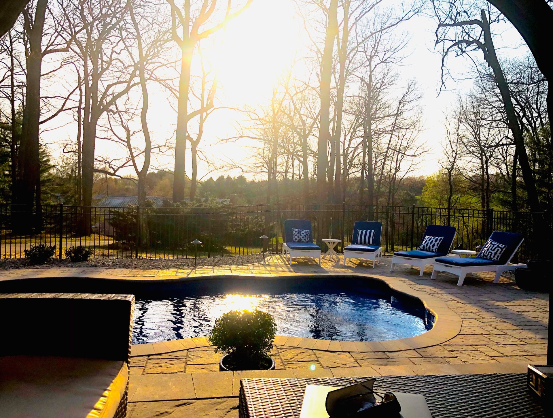 A sunlit swimming pool surrounded by a patio with lounge chairs, overlooking trees at sunset.