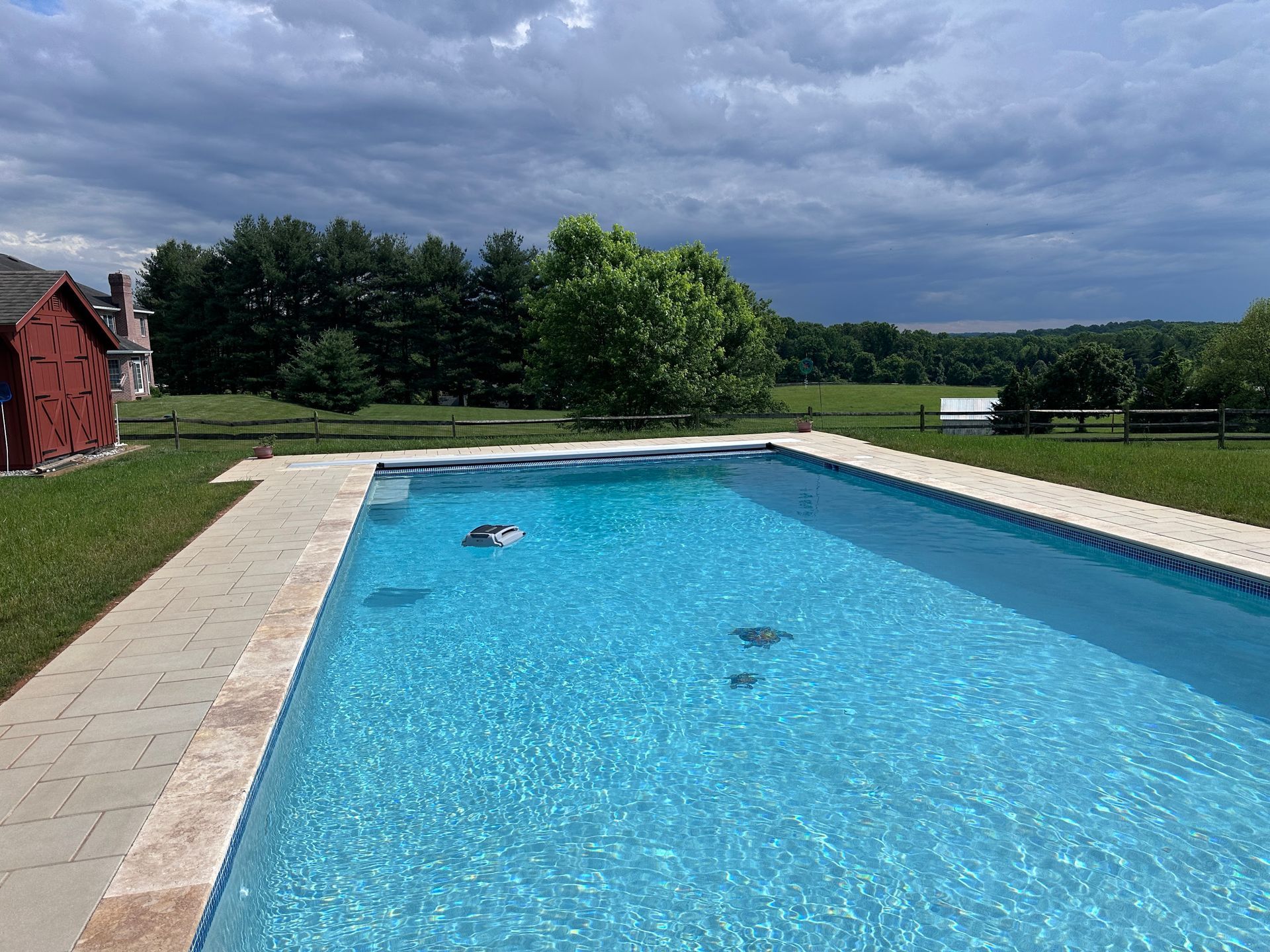 A swimming pool in a grassy yard under a cloudy sky, with a red building to the side and trees in the background.