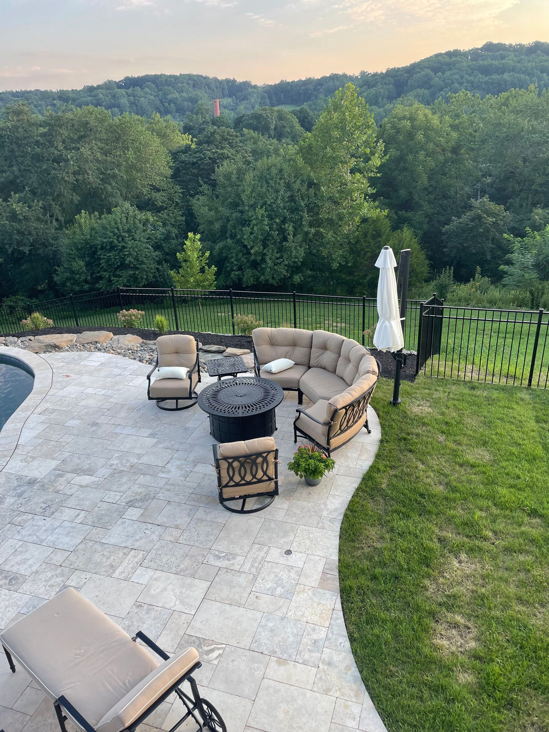 Outdoor patio with stone pavers, a curved seating area, fire pit, and lounge chairs overlooking a wooded mountain view.