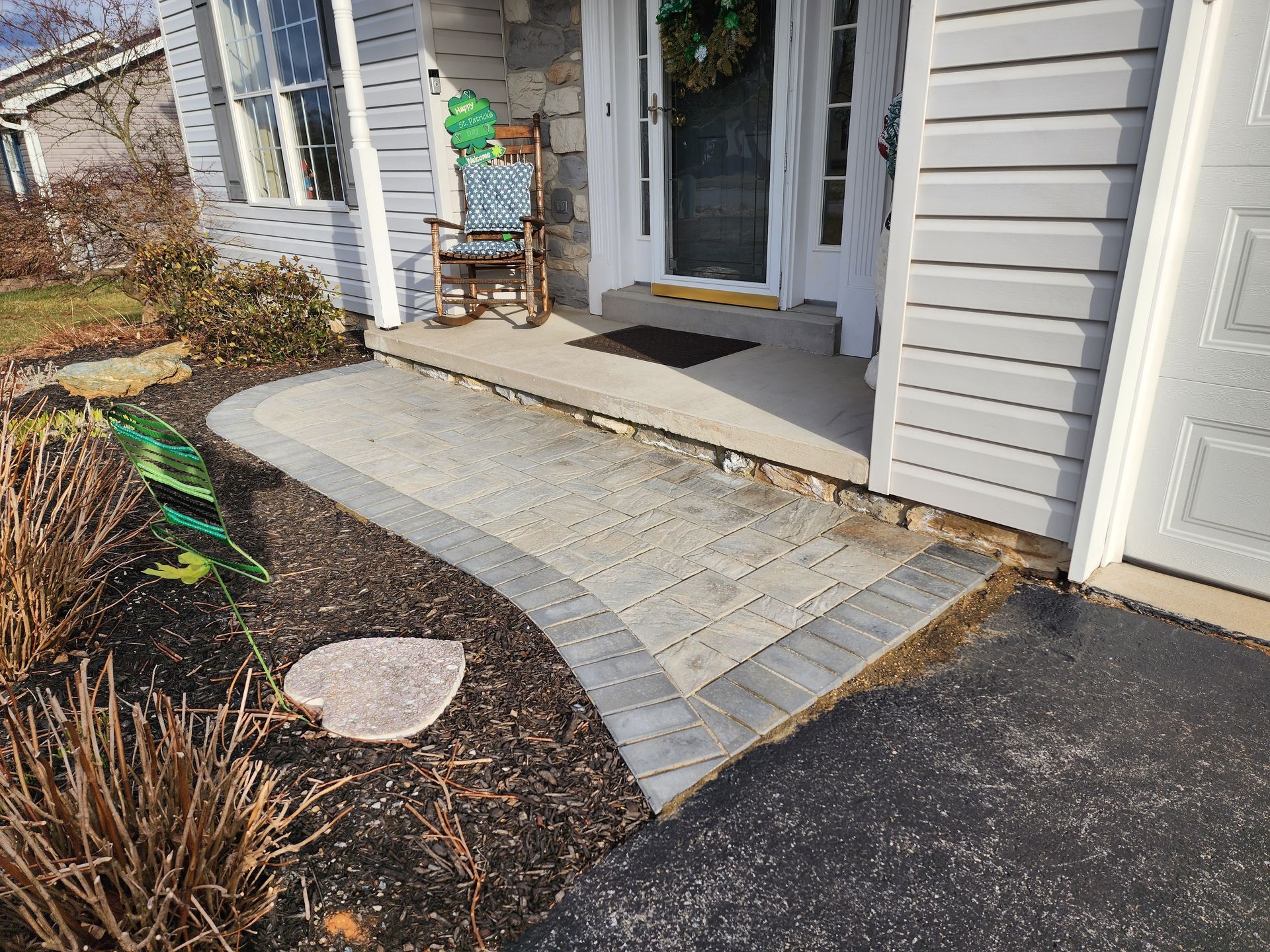 A paver walkway leads to a front porch and entryway of a home, with a rocking chair visible near the front door.