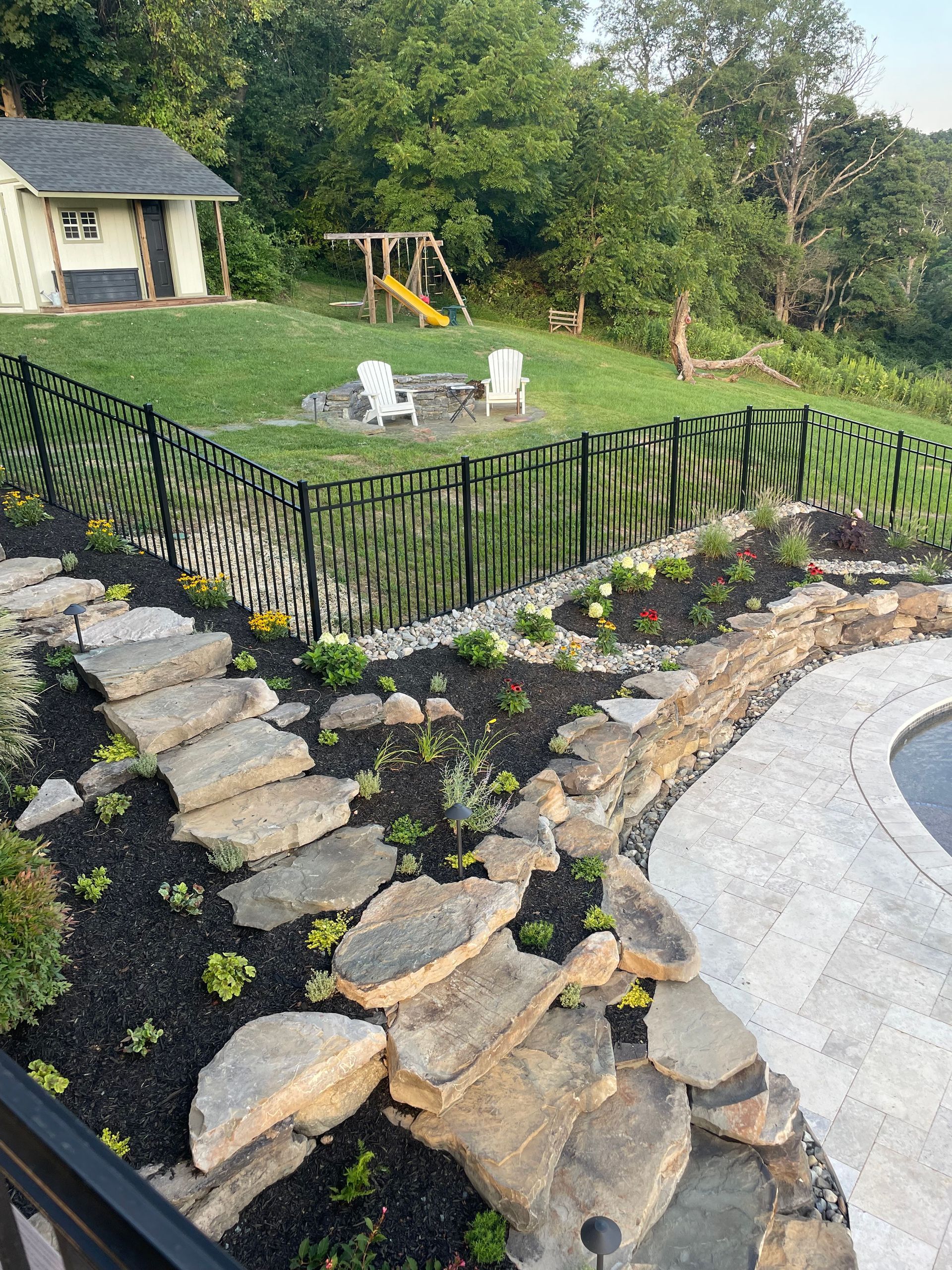 A stone stairway leads through a landscaped garden near a swimming pool, overlooking a yard with a playset and shed.