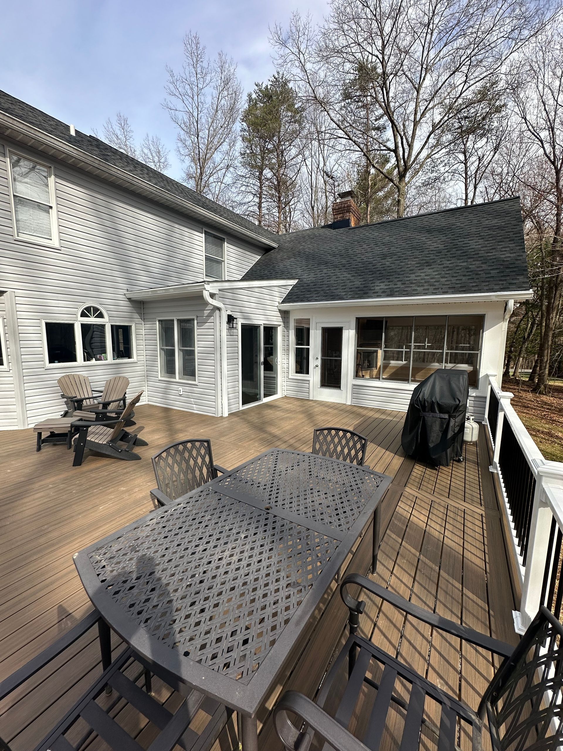 A wooden deck with a patio dining set, seating, and a grill attached to a multi-story home with a screened-in porch.