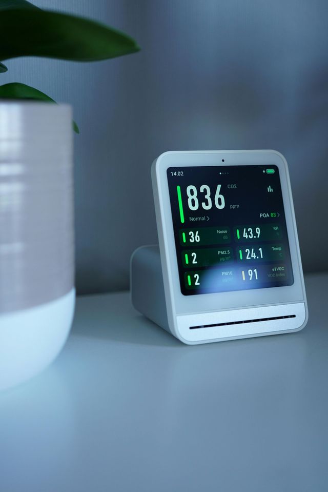 A smart air quality monitor showing numerical data on its screen, placed on a white surface next to a houseplant.