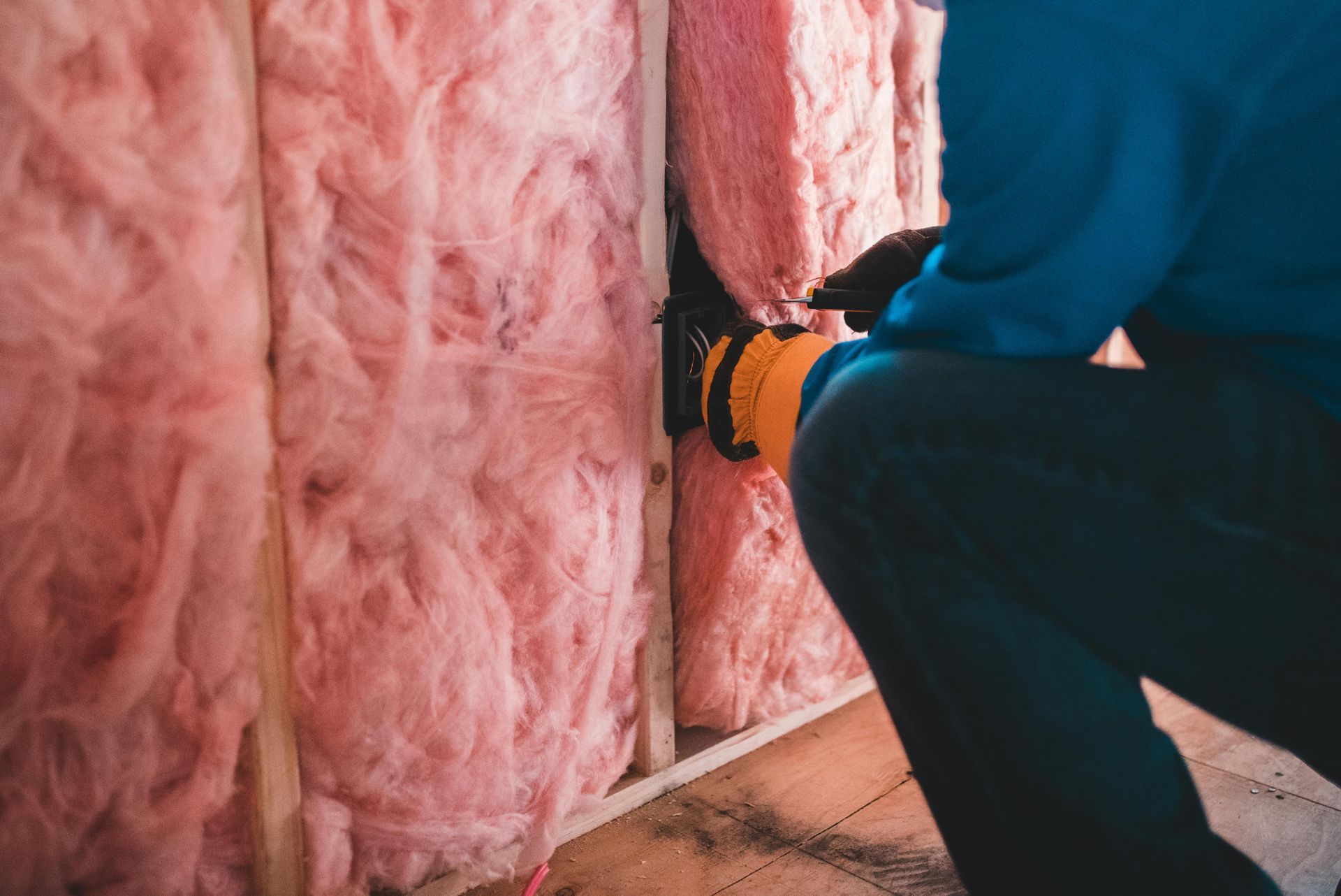 A person in a blue shirt and yellow gloves installs pink fiberglass insulation between wooden wall studs.