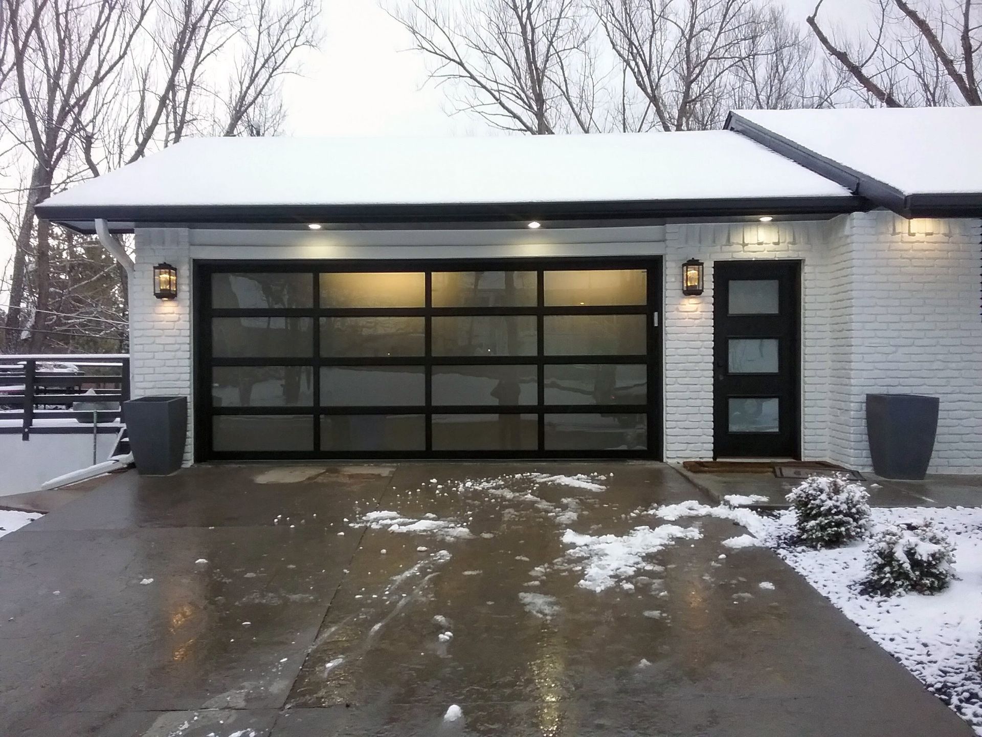 Modern garage with glass door and black trim, snowy driveway and roof, and exterior lighting.