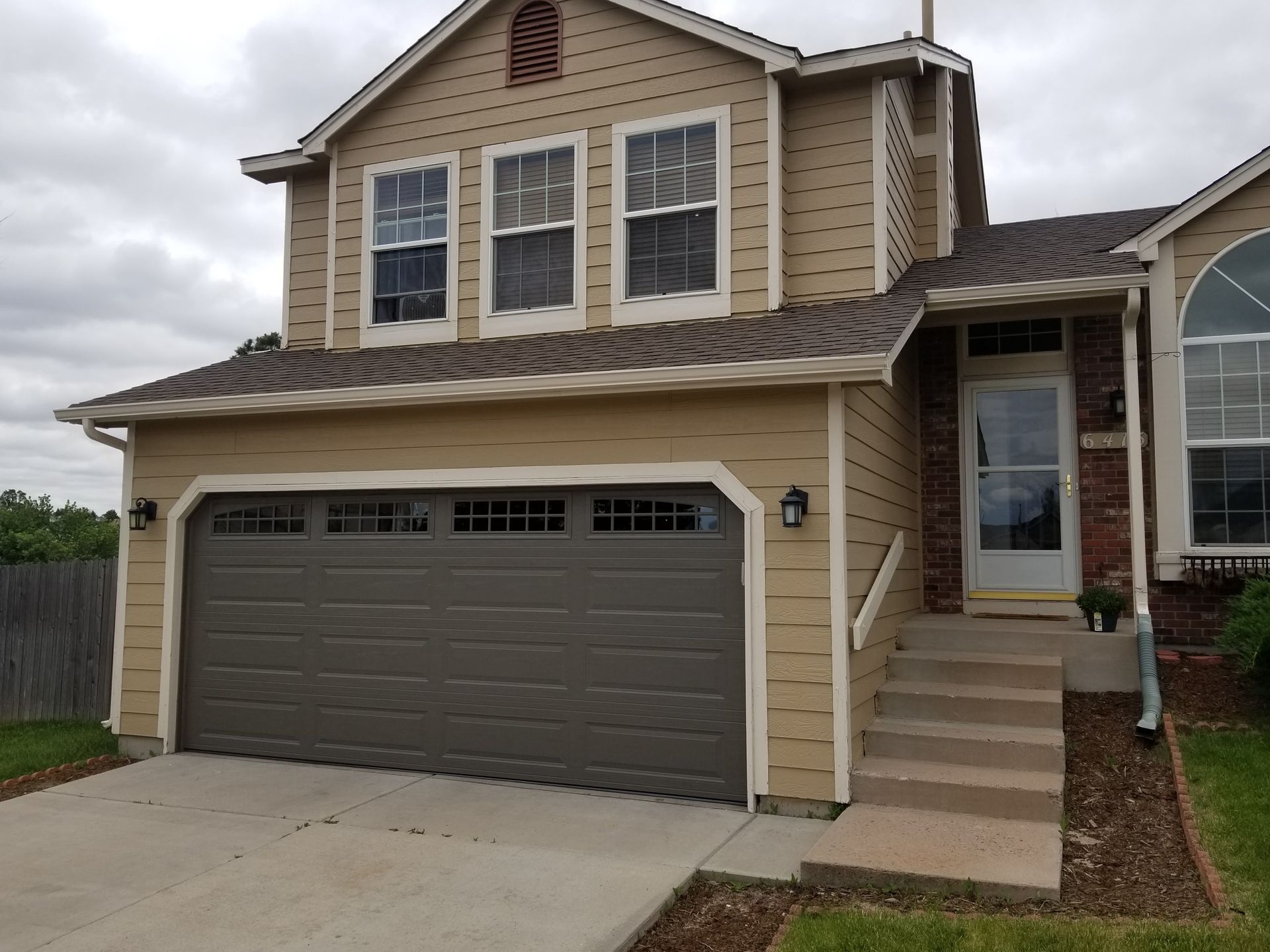 Tan two-story house with a grey garage door, three windows upstairs, and a front door with steps.