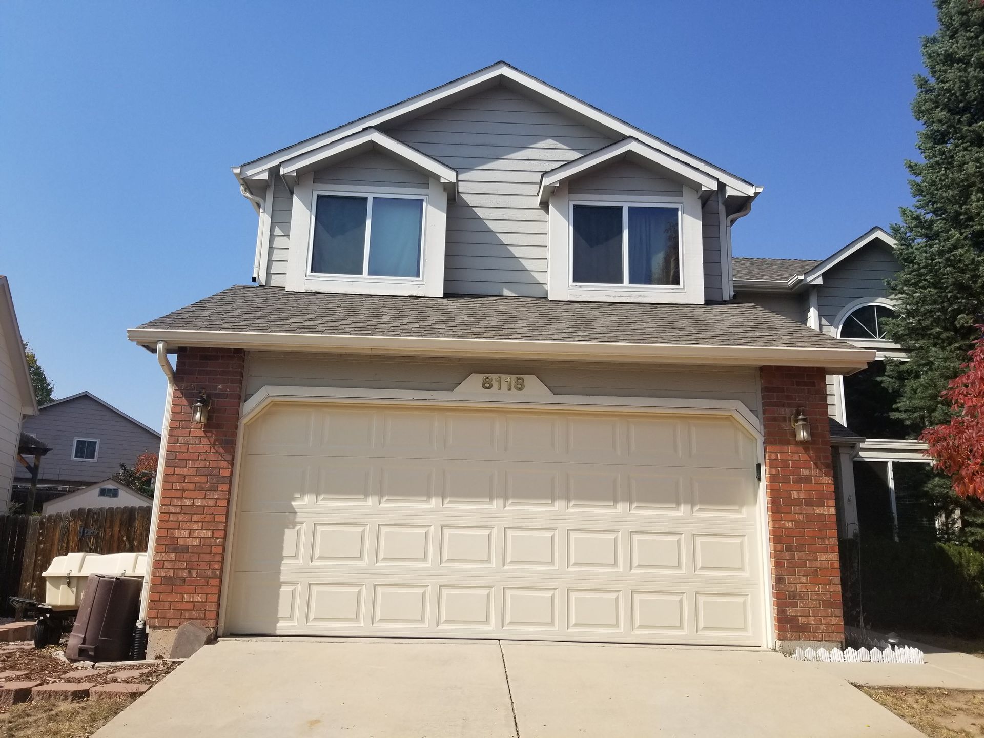 Two-story house with beige garage door, red brick accents, gray siding, and two dormer windows under a clear blue sky.