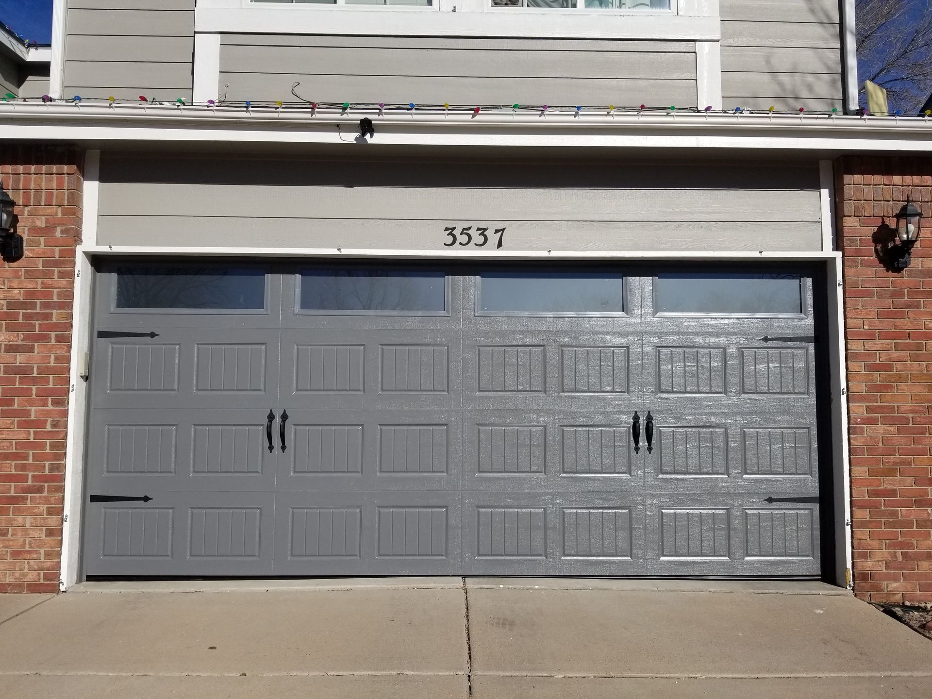 Gray garage door with decorative panels, windows, and black handles; brick and siding exterior.