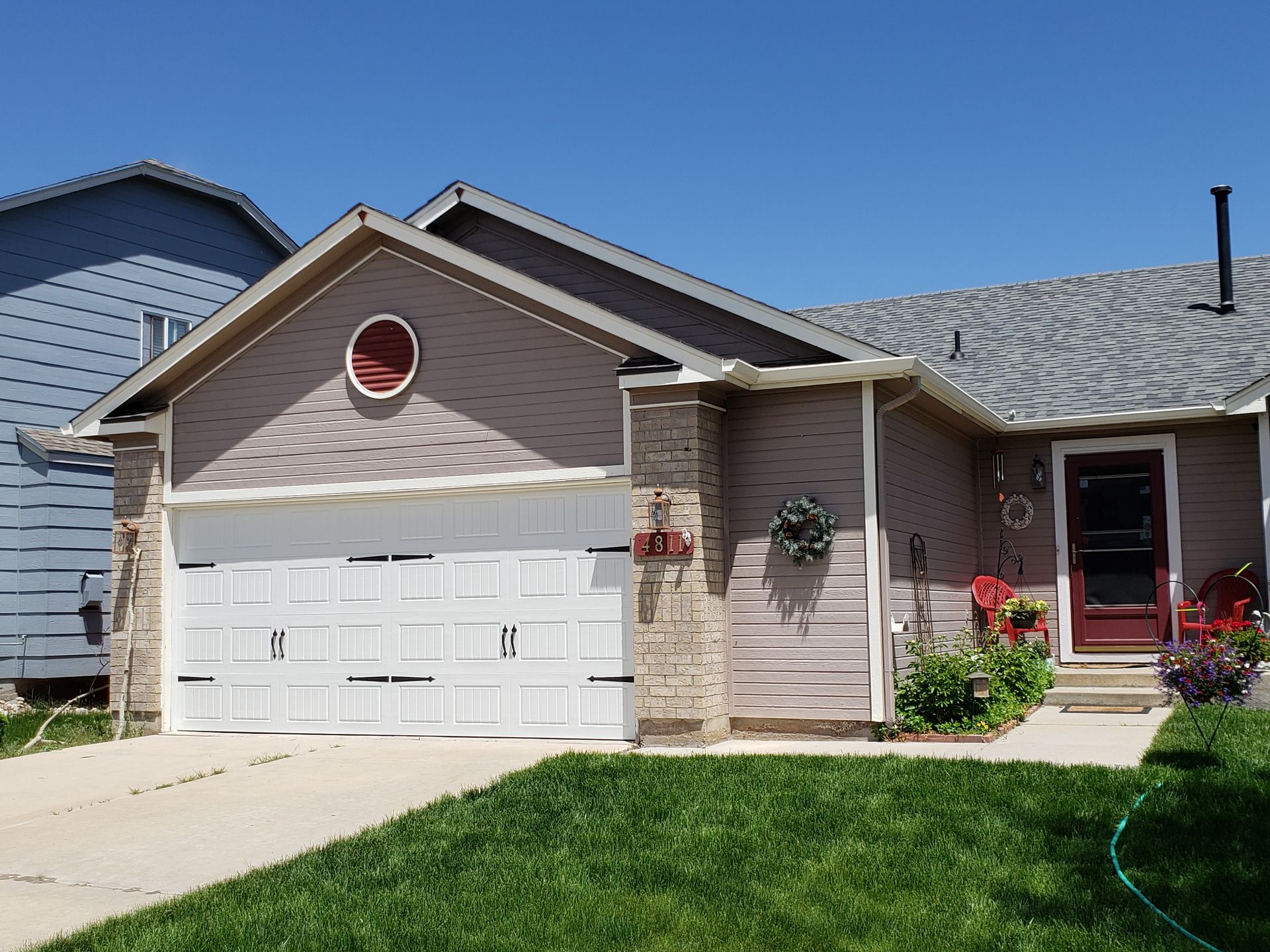 Tan and gray house with a white garage door, red door, and green lawn on a sunny day.