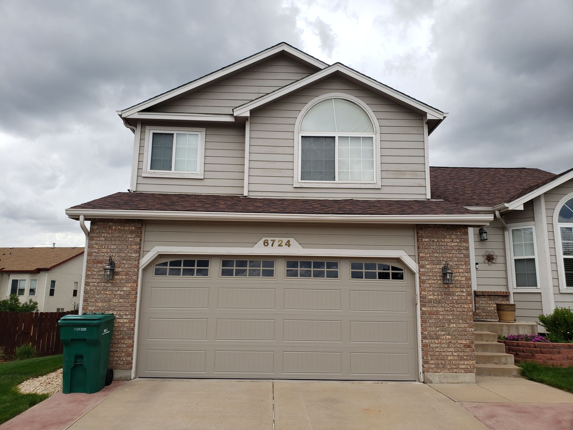 Two-story house with tan siding and brown roof. Beige garage door, stone columns, and cloudy sky.