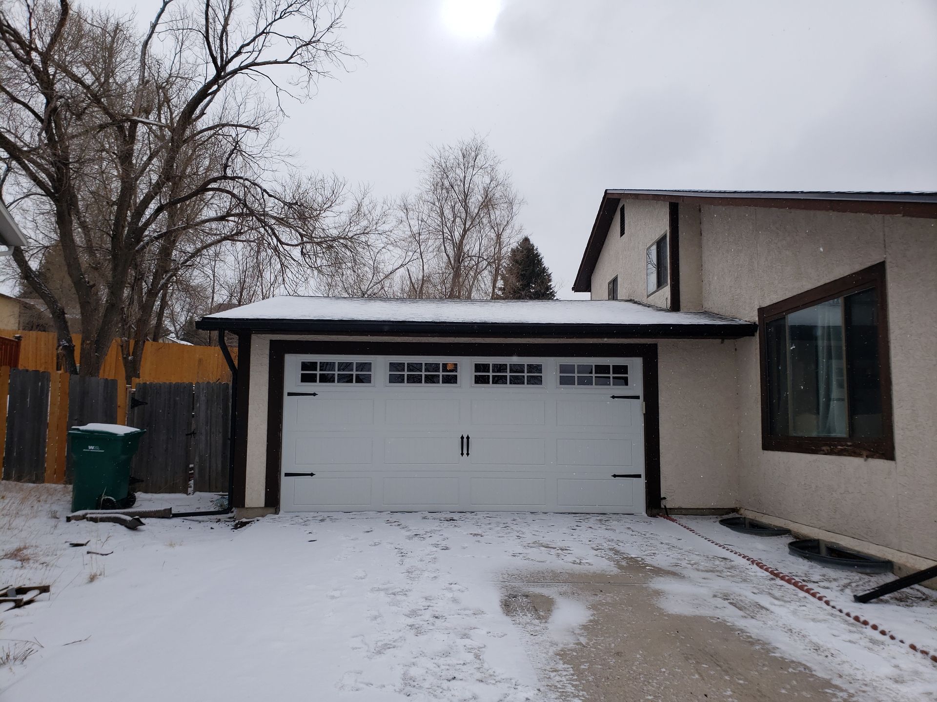 Snowy scene: white garage door, dark trim, and a house with a window. The driveway and ground are covered in snow.