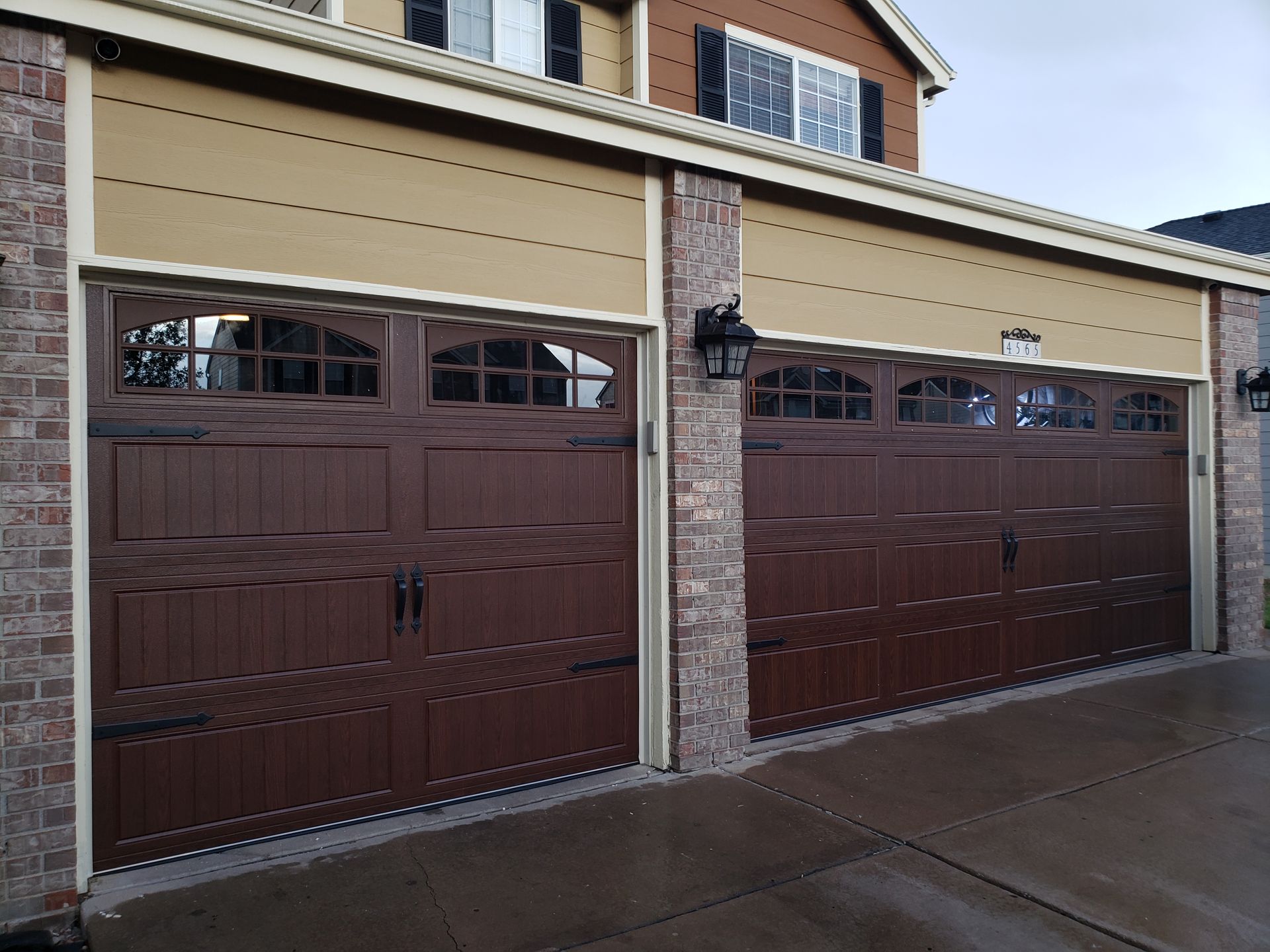 Two brown garage doors with decorative windows on a brick and tan house.