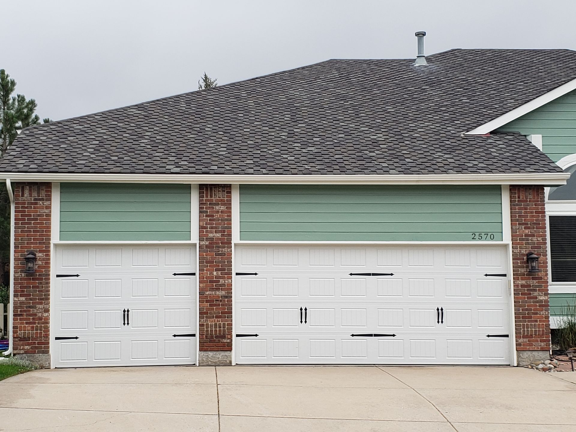 White garage doors with red brick columns, green siding, gray roof, and a concrete driveway.
