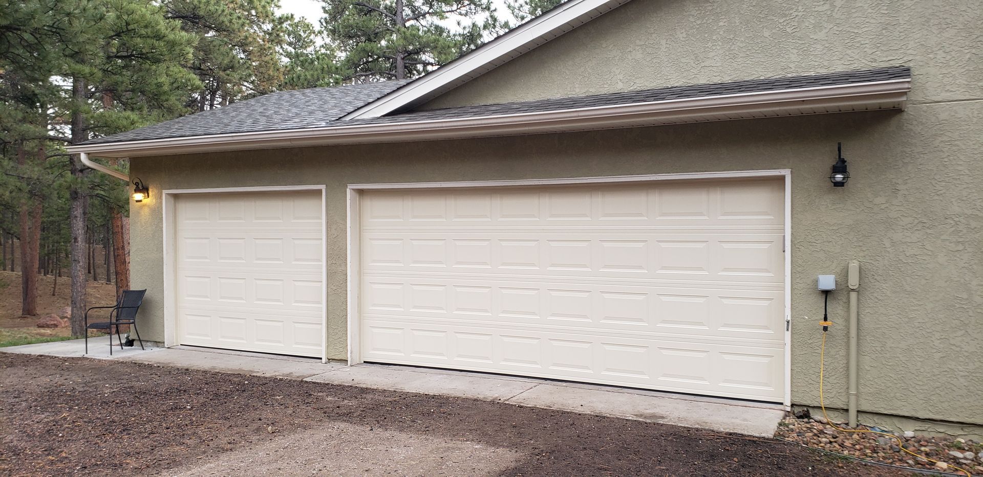 Garage with two white doors, tan stucco exterior, gray roof, and surrounding trees.