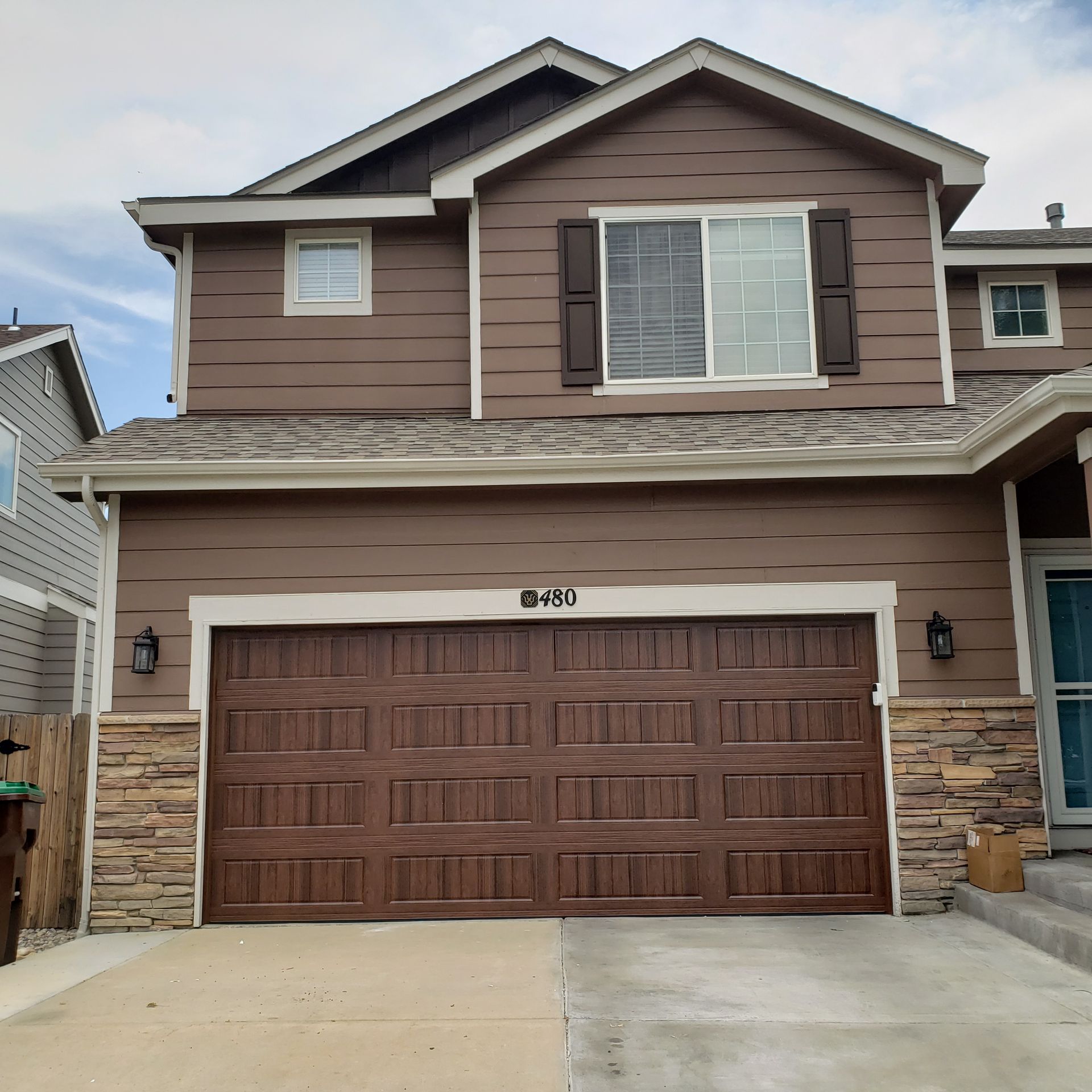 Brown two-story house with a matching garage door, stone accents, and a concrete driveway on a sunny day.