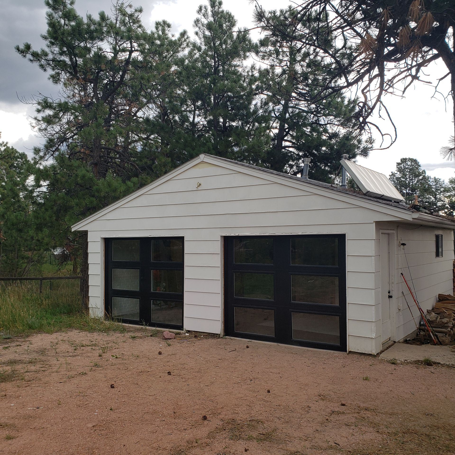 White garage with black garage doors, in a dirt yard with trees.