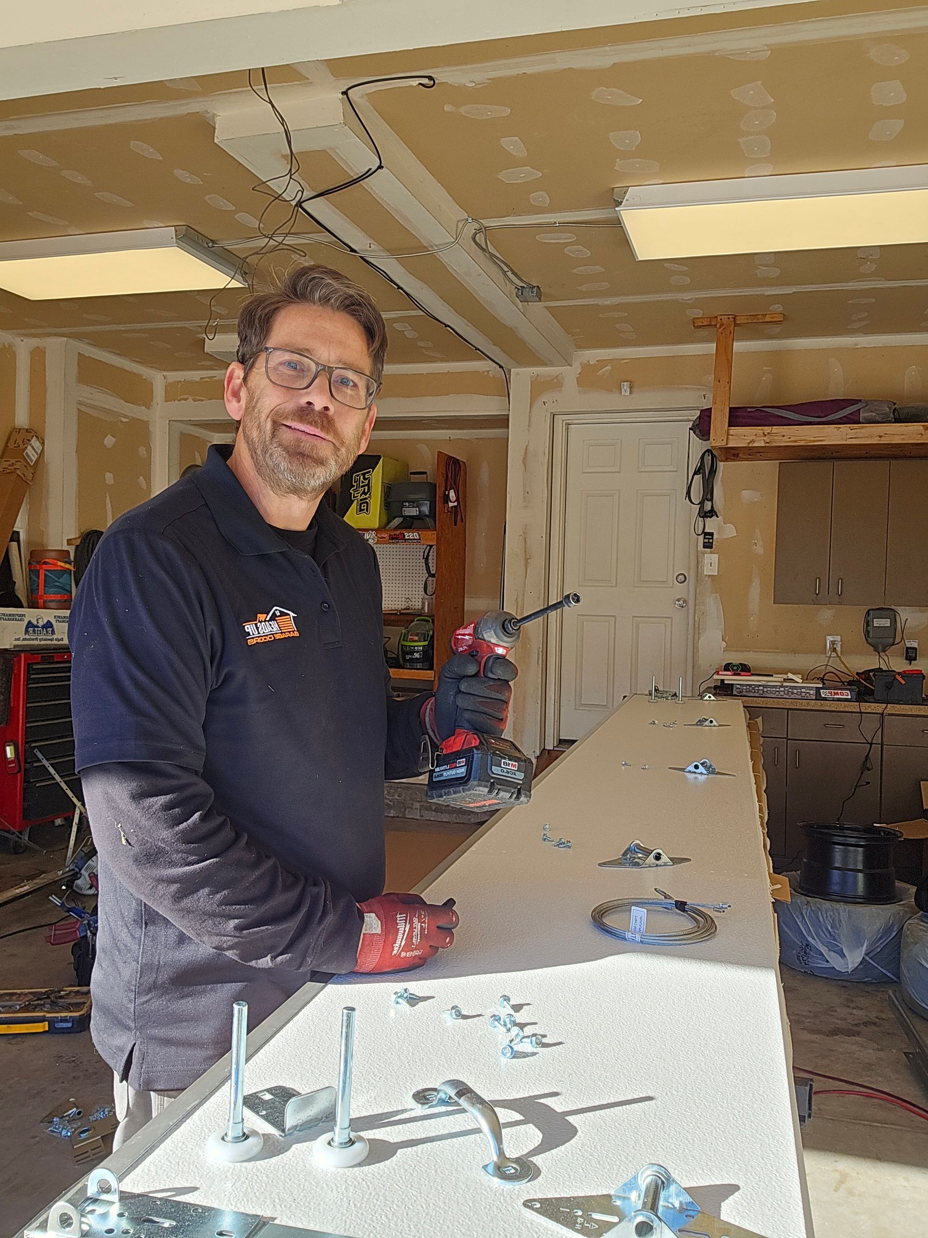 Man in a garage holding a drill, looking at the camera. He's standing by a table with tools and objects on it.
