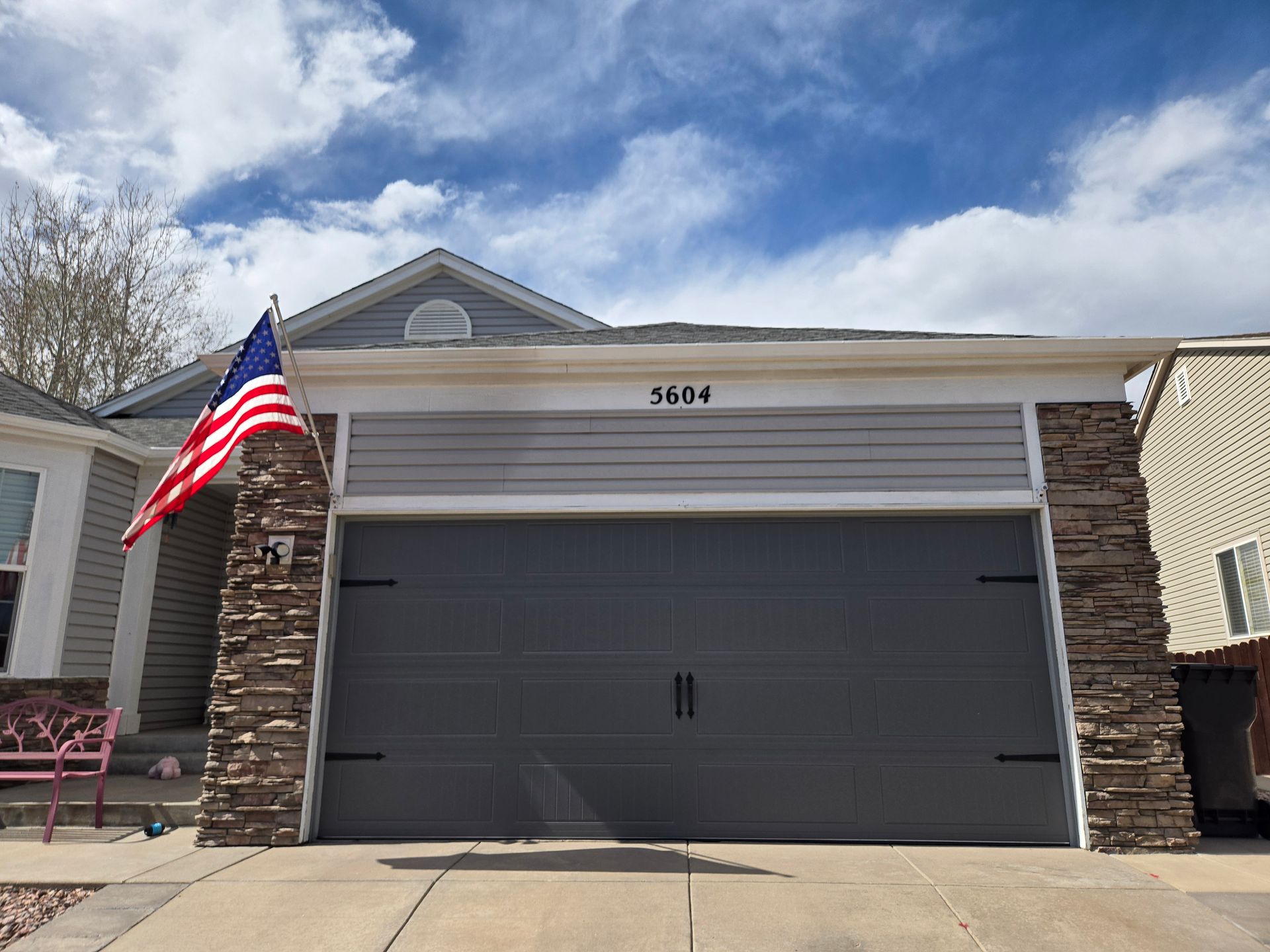 A gray garage door with an American flag, house number 4614, and stone columns. Blue sky.