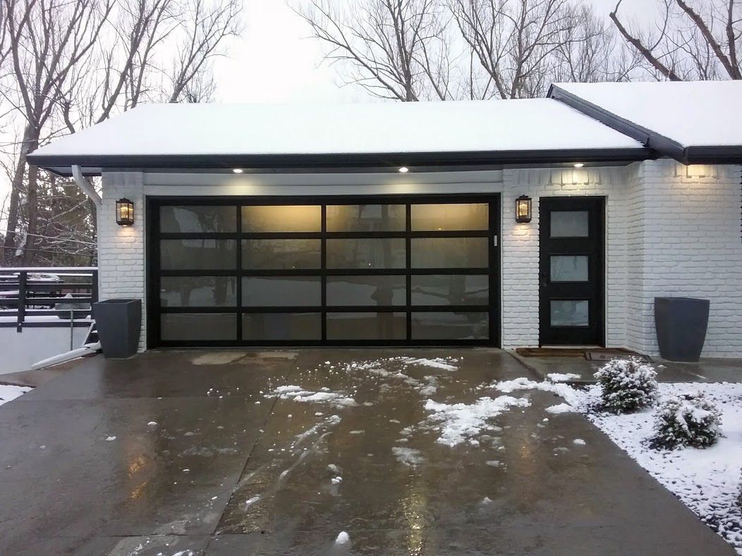 Garage with black-framed glass door, brick exterior, and snow-covered driveway. Two sconces and a front door.