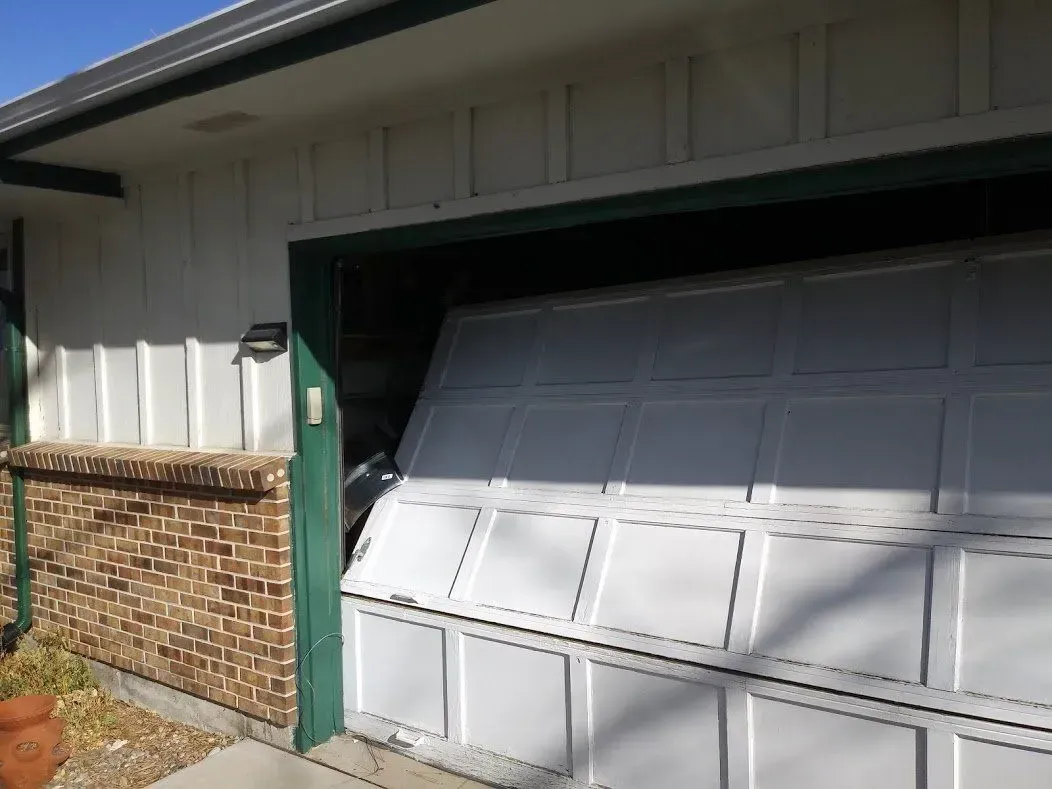 White garage door partially open, appearing damaged. Brick and white siding on exterior.
