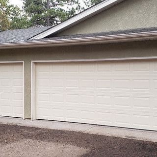 Two-car garage with off-white doors and light green exterior. Asphalt driveway, trees in the background.