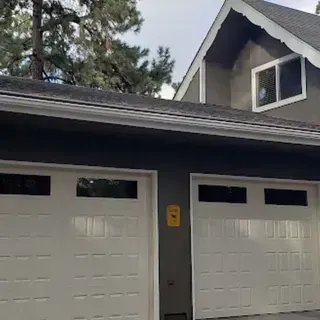 Two white garage doors with black-framed windows, against a gray house exterior, with trees and sky.