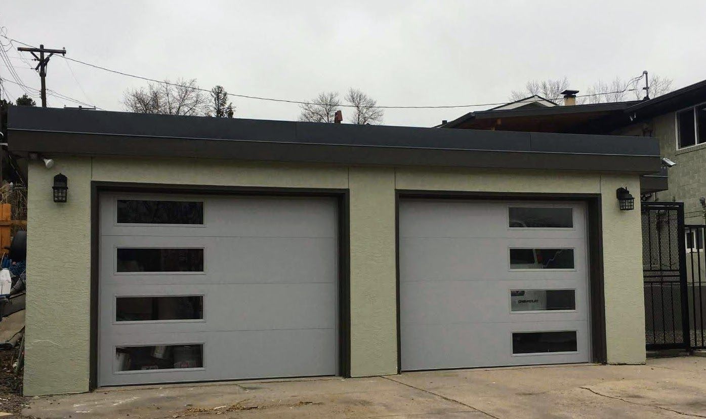 Two-car garage with gray doors, windows, and light tan stucco exterior. Flat roof.