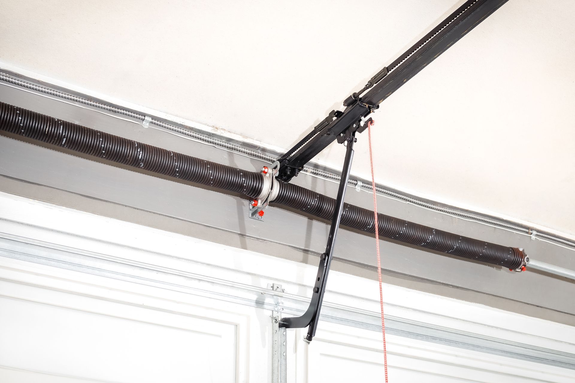 Garage door opener mechanism with a coiled spring and track on a white ceiling.