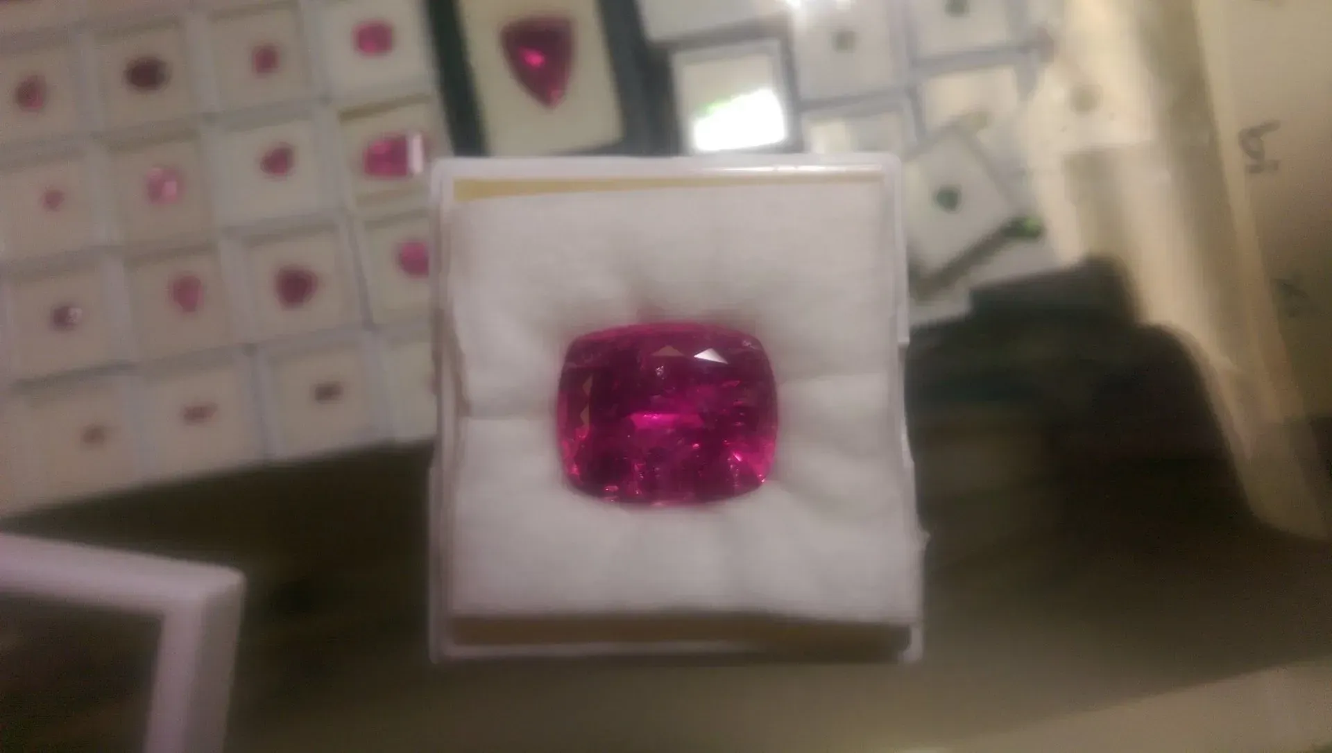 A small pink gemstone in a white display box, with blurred jewelry boxes in the background.