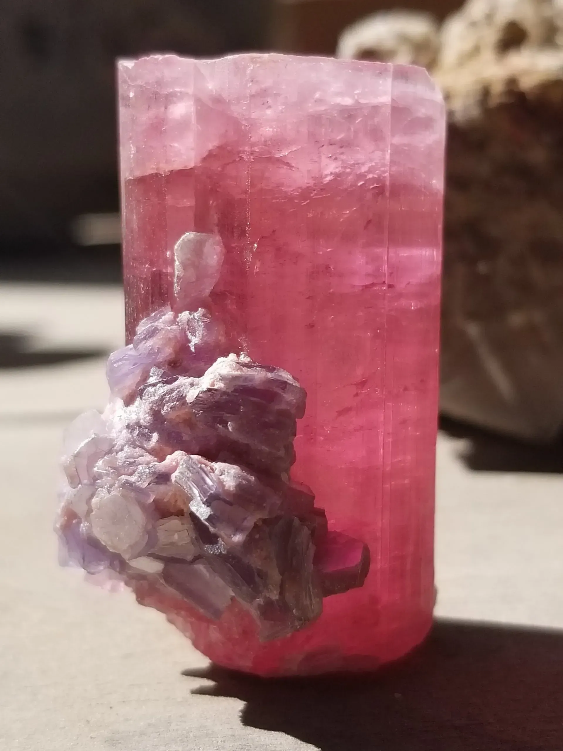 Pink crystal with a purple mineral cluster attached on a light surface