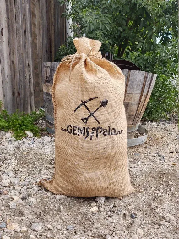 Burlap sack with “GEMS PALA” logo standing on gravel outdoors beside wood and plants