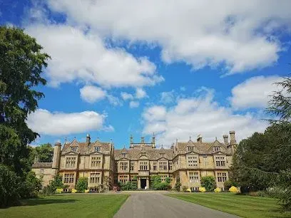 Large stone manor house under a blue sky with fluffy white clouds, with a long driveway.