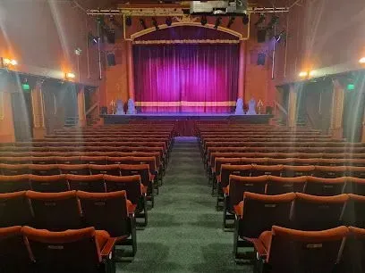 Inside a theater: empty red seating faces a stage with purple curtains.