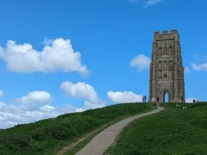 Stone tower on green hill with path, blue sky, and white clouds.
