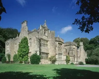 Stone manor house with multi-gabled roof and mullioned windows, set on green lawn under blue sky.