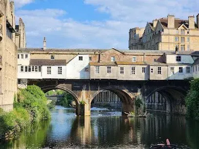 Pulteney Bridge in Bath, England, with shops built along it. Arches over the river, stone buildings, blue sky.
