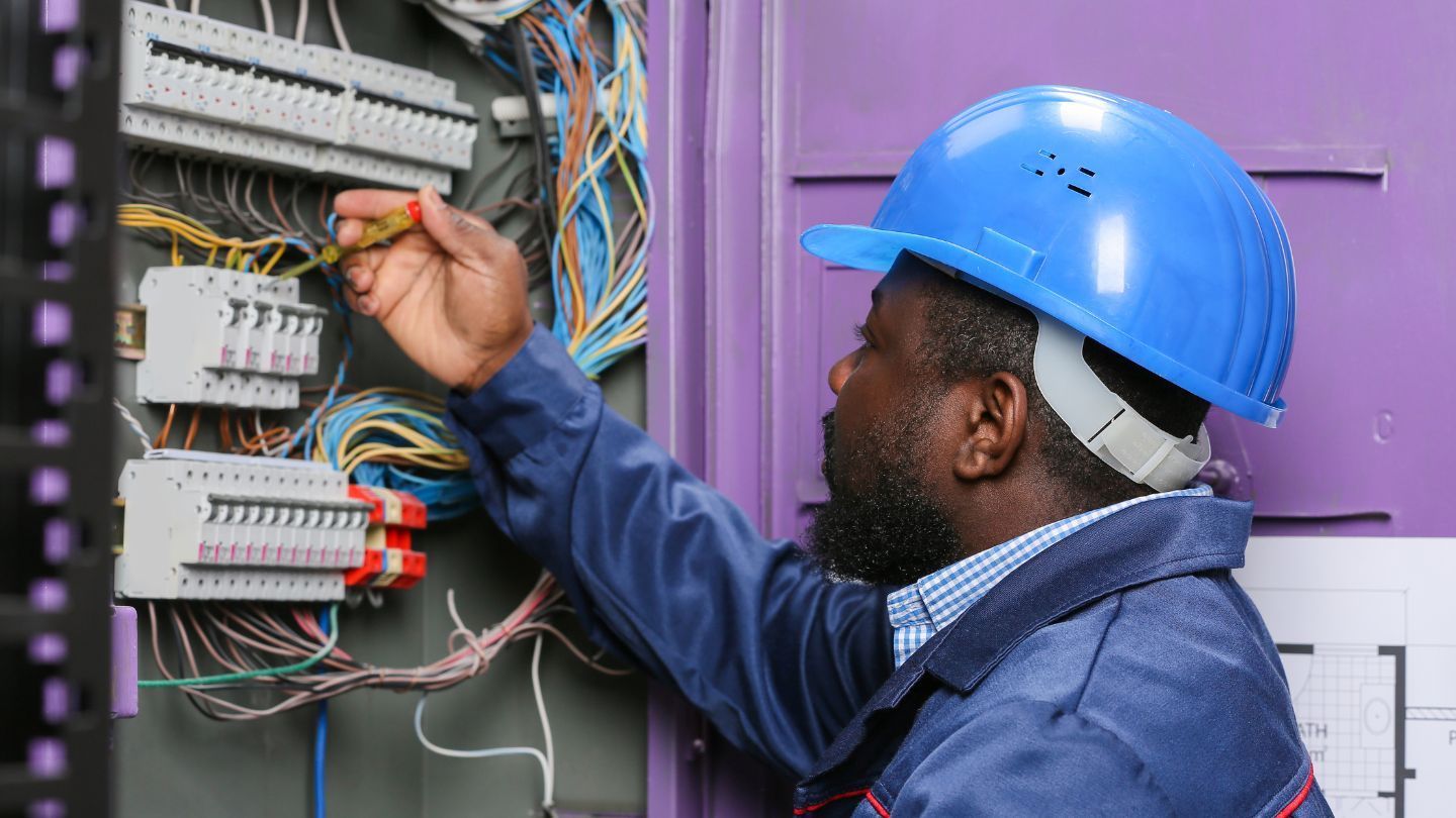 Roofers installing shingles on a house roof. One hammers, the other works in the background. Blue sky overhead.