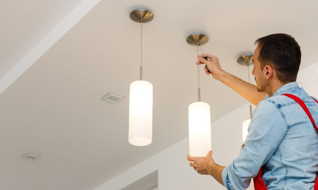 Electrician installing a pendant light, standing on a ladder in a white room.