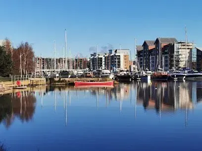 Harbor with boats and buildings reflecting in calm blue water under a clear sky.