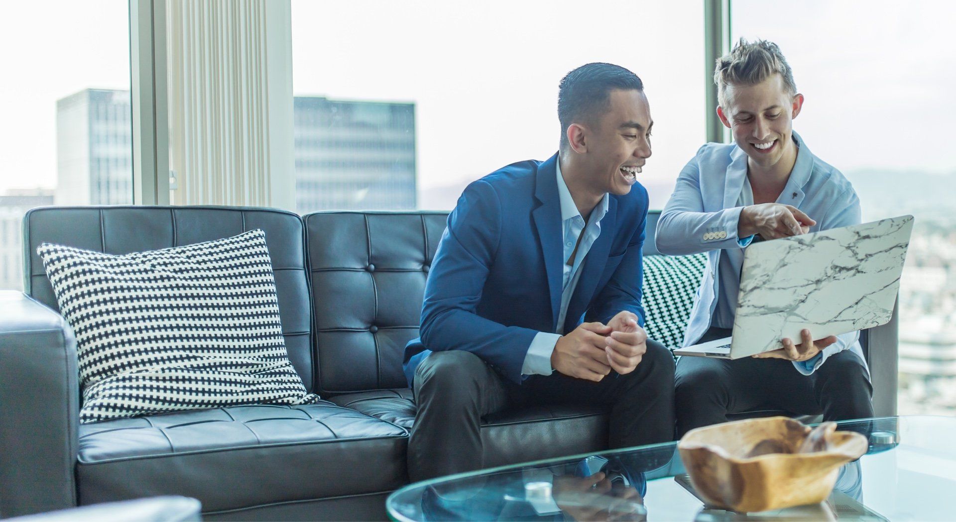 Two men in suits looking at laptop, smiling. They sit on a black couch in a modern office, overlooking city.
