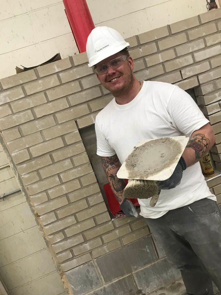 A man wearing a hard hat and holding a trowel is standing in front of a brick wall.