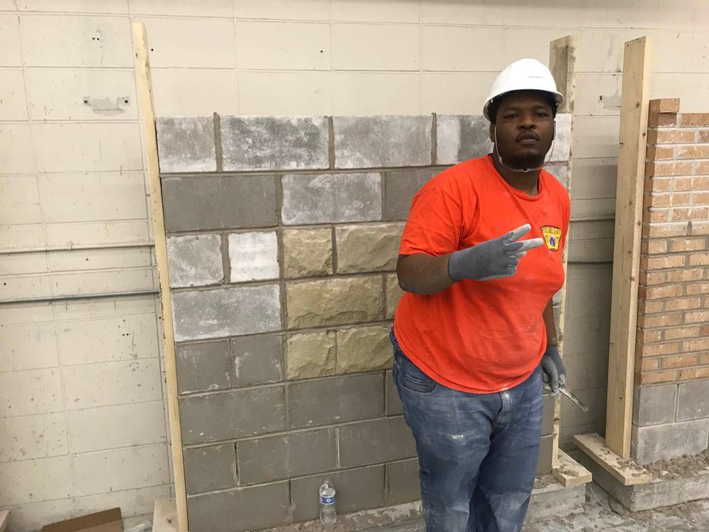 A man wearing a hard hat and an orange shirt is standing in front of a brick wall.