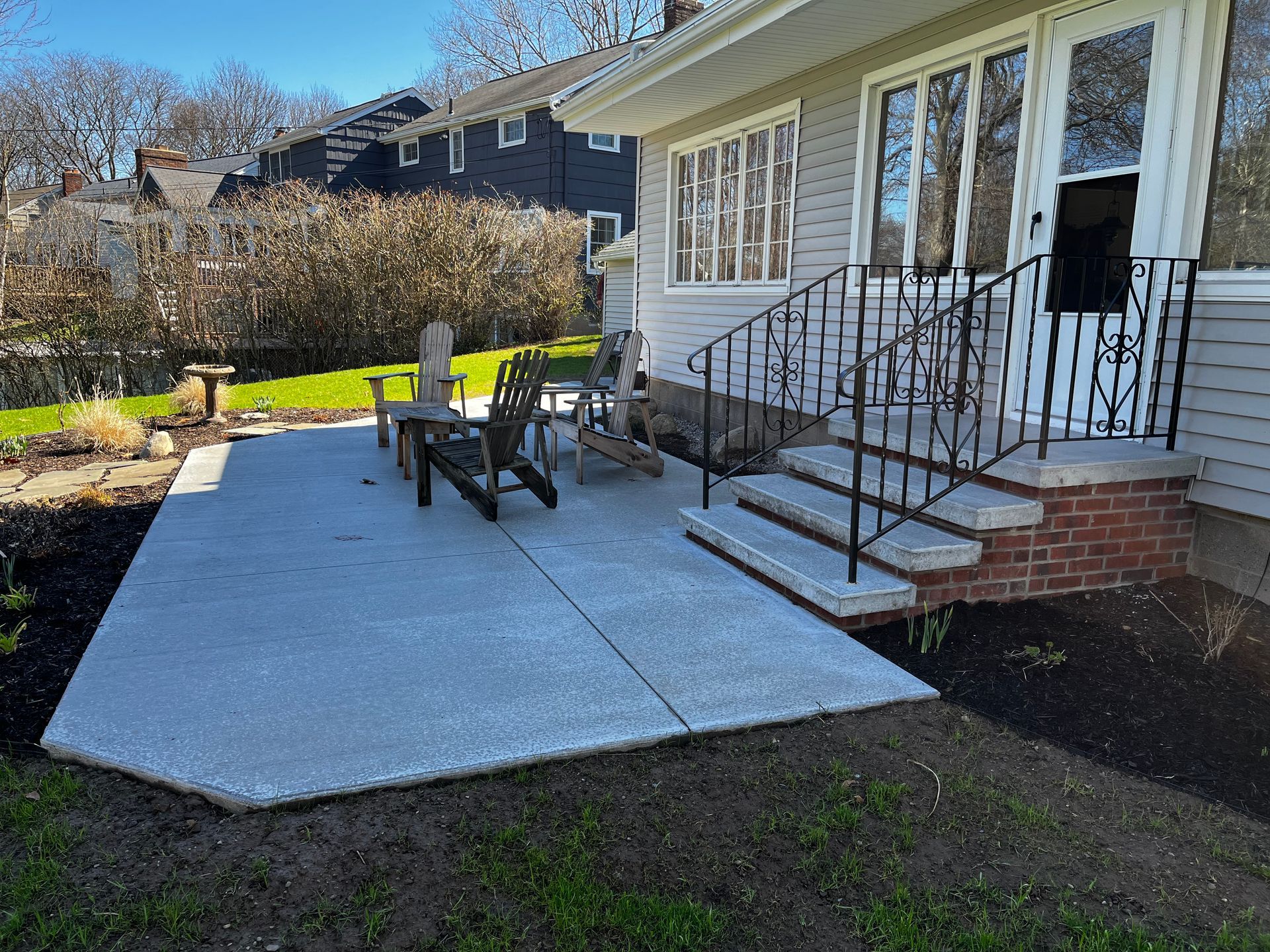 A concrete patio with two wooden chairs sits beside a house with brick steps and black iron railings.